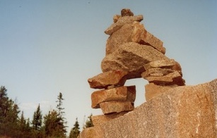 A stone Inukshuk against a clear sky with evergreens in the background