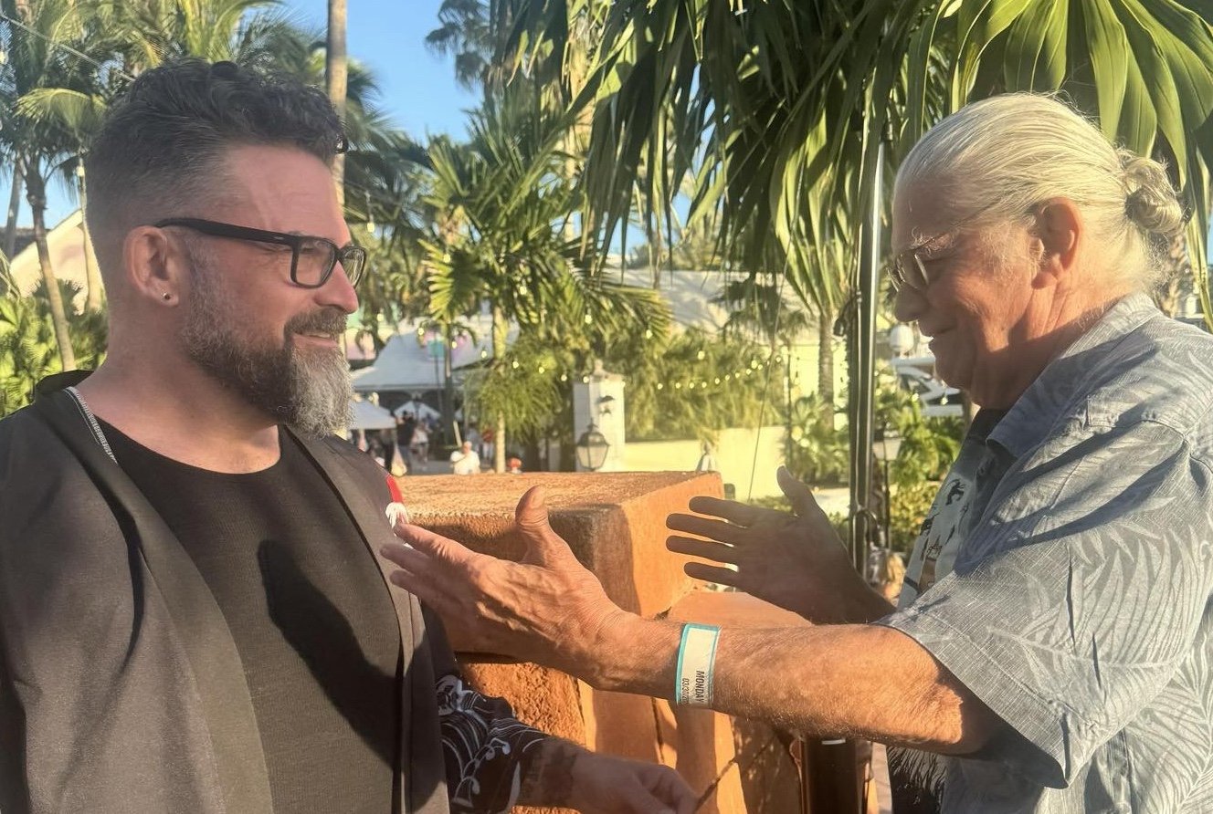 Ross and Thomas reaching toward each other for a hug, palm trees in the background